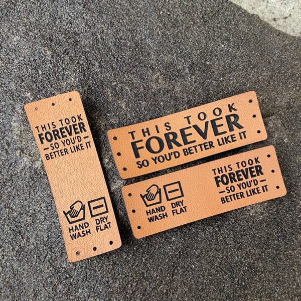 Mixed set of six faux leather tags with 'This took forever so you’d better like it' text. Includes two brim fold, two side-fold, and two flat stitch tags. Available in cinnamon brown or warm gray.